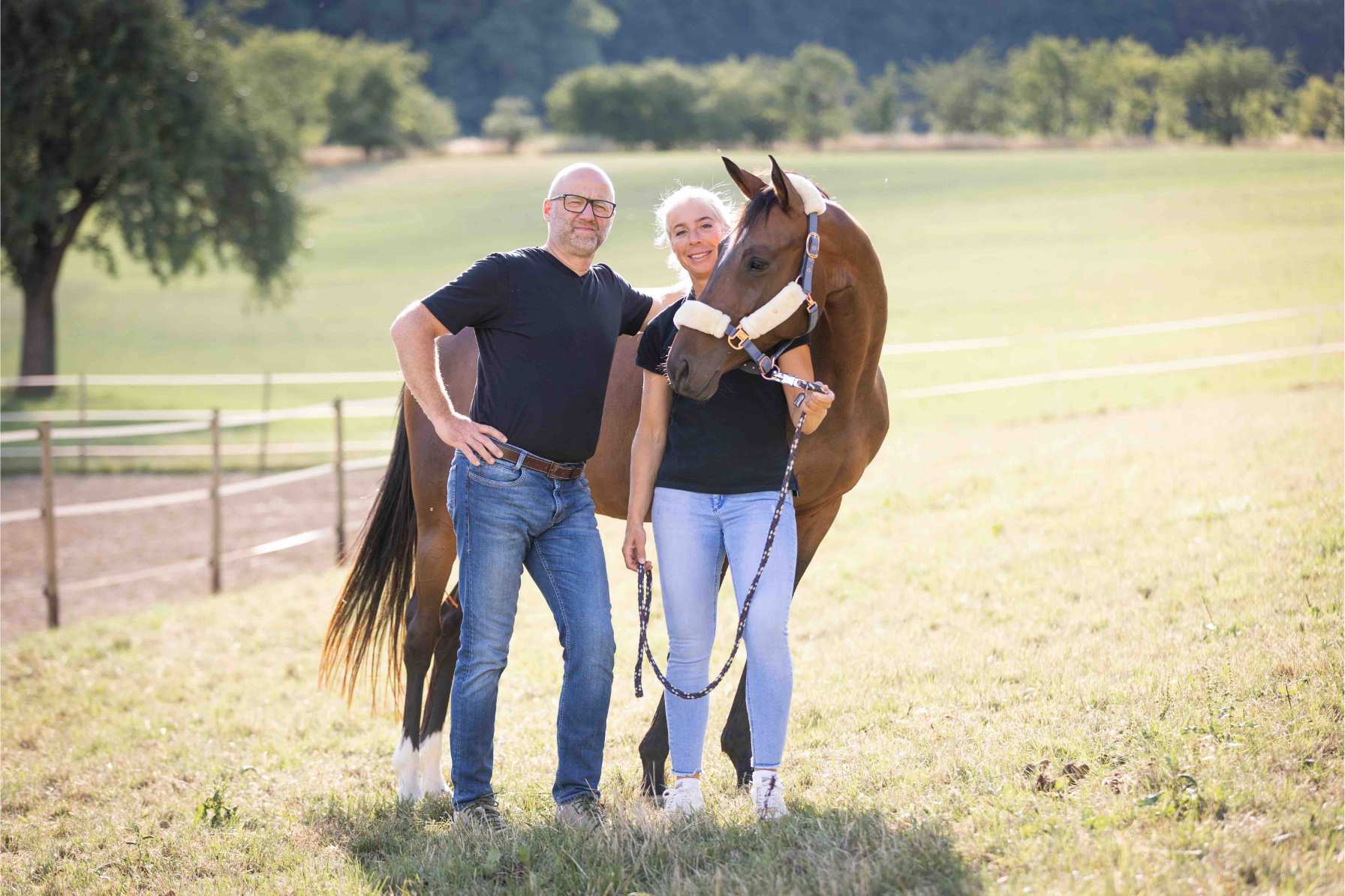 Heiner und Anja Beifuss stehen zusammen mit einem braunen Pferd auf einer Weide bei sehr hellem Sonnenlicht