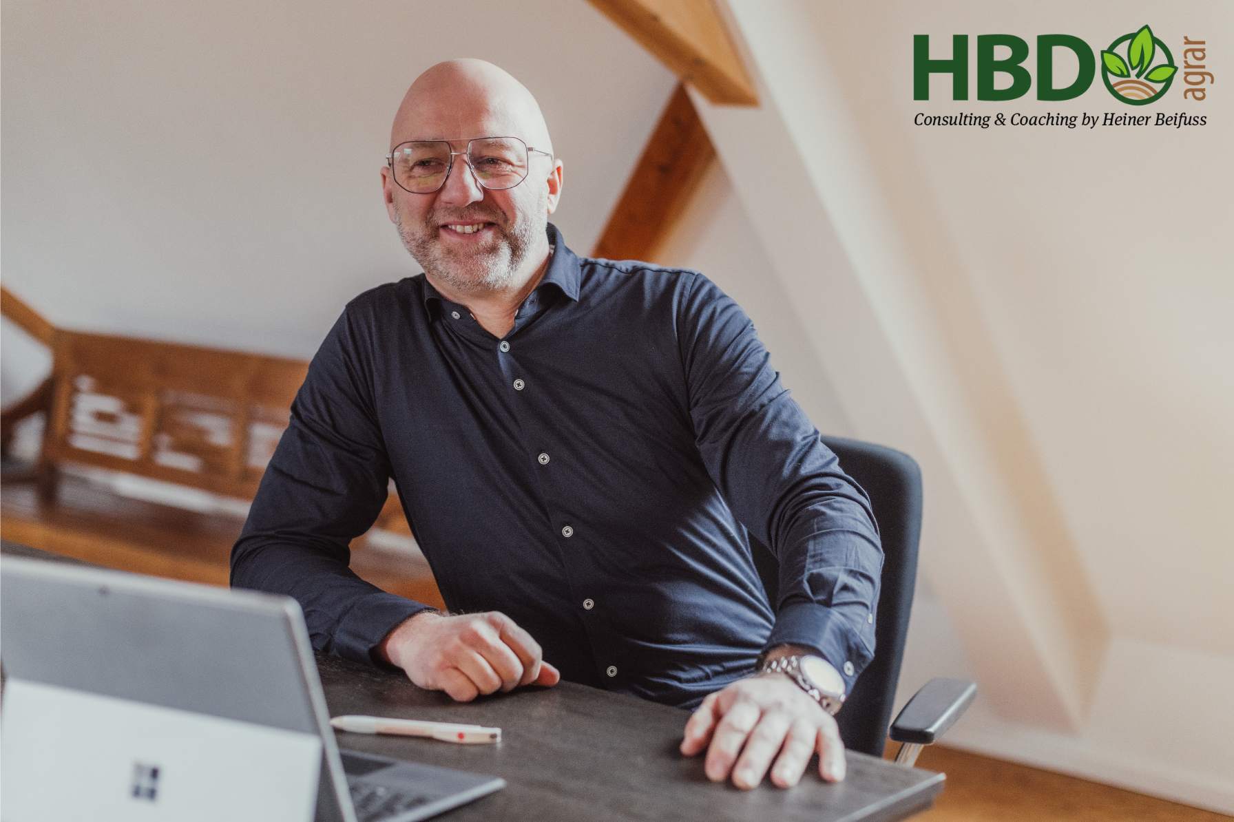 Heiner Beifuss sitzt an seinem Schreibtisch vor seinem Laptop in einem Raum mit Dachbalken. Außerdem ist das HBD Agrar - Consulting und Coaching by Heiner Beifuss - Logo zu sehen